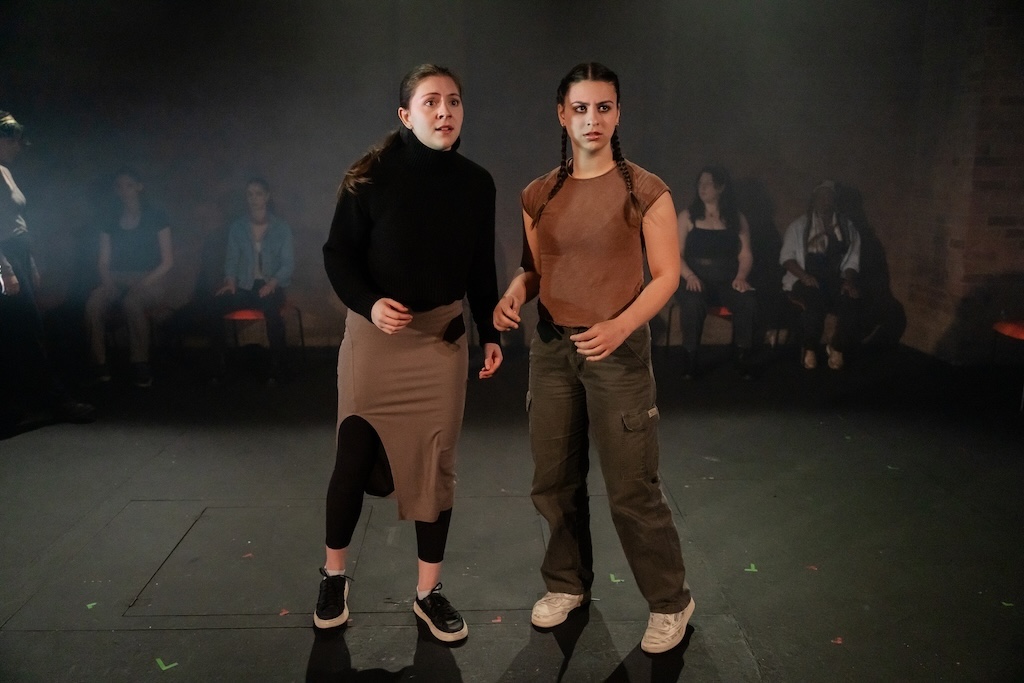 Action shot of the spring 2024 Shakespeare in Performance at RADA final presentation of Julius Caesar. Two actors stand on stage in mid-performance, facing the camera while other seated actors look on.